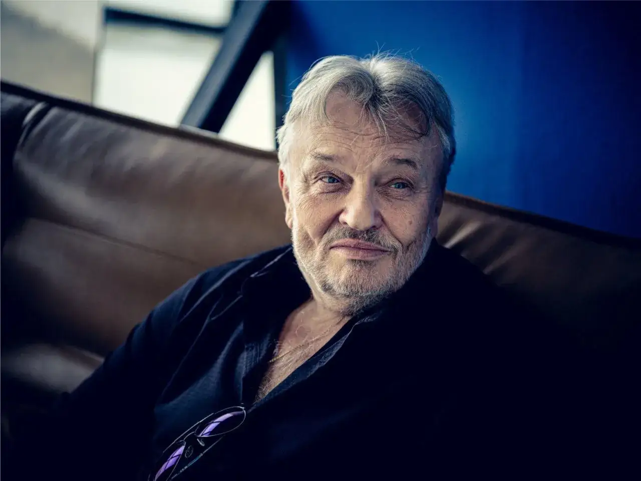Uśmiechnięty Krzysztof Cugowski, siedzący na kanapie. Wiek artysty to 69 lat.