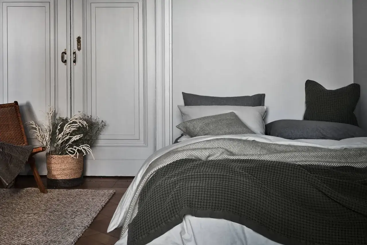 Gem&uuml;tliche Bettw&auml;sche in Grau- und Gr&uuml;nt&ouml;nen, inspiriert von modernen Bett machen Ideen. Ein Korb mit Trockenblumen und ein Sessel vervollst&auml;ndigen die Szene.