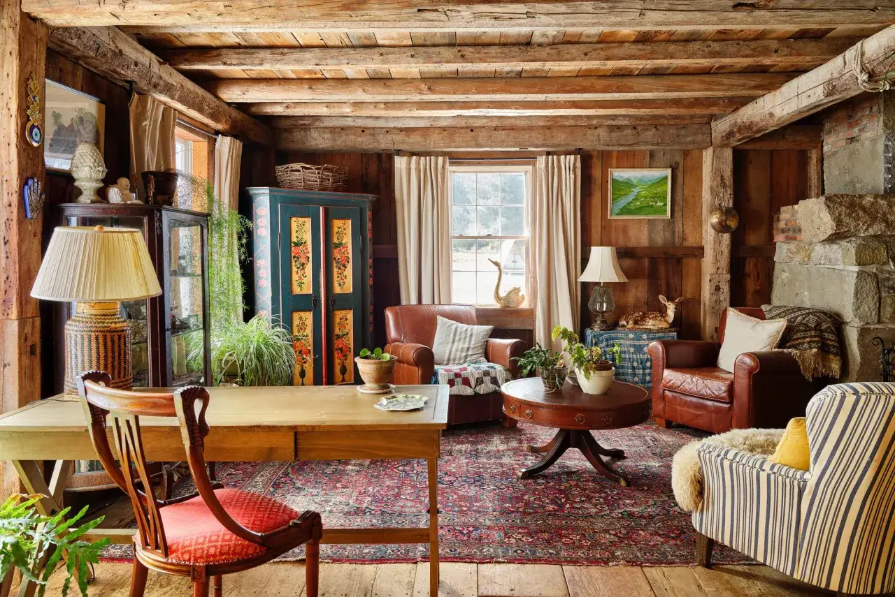 Cozy cabin living room with a rustic, cottagecore aesthetic. Features a wooden desk, leather armchairs, a floral cabinet, and a vintage rug.