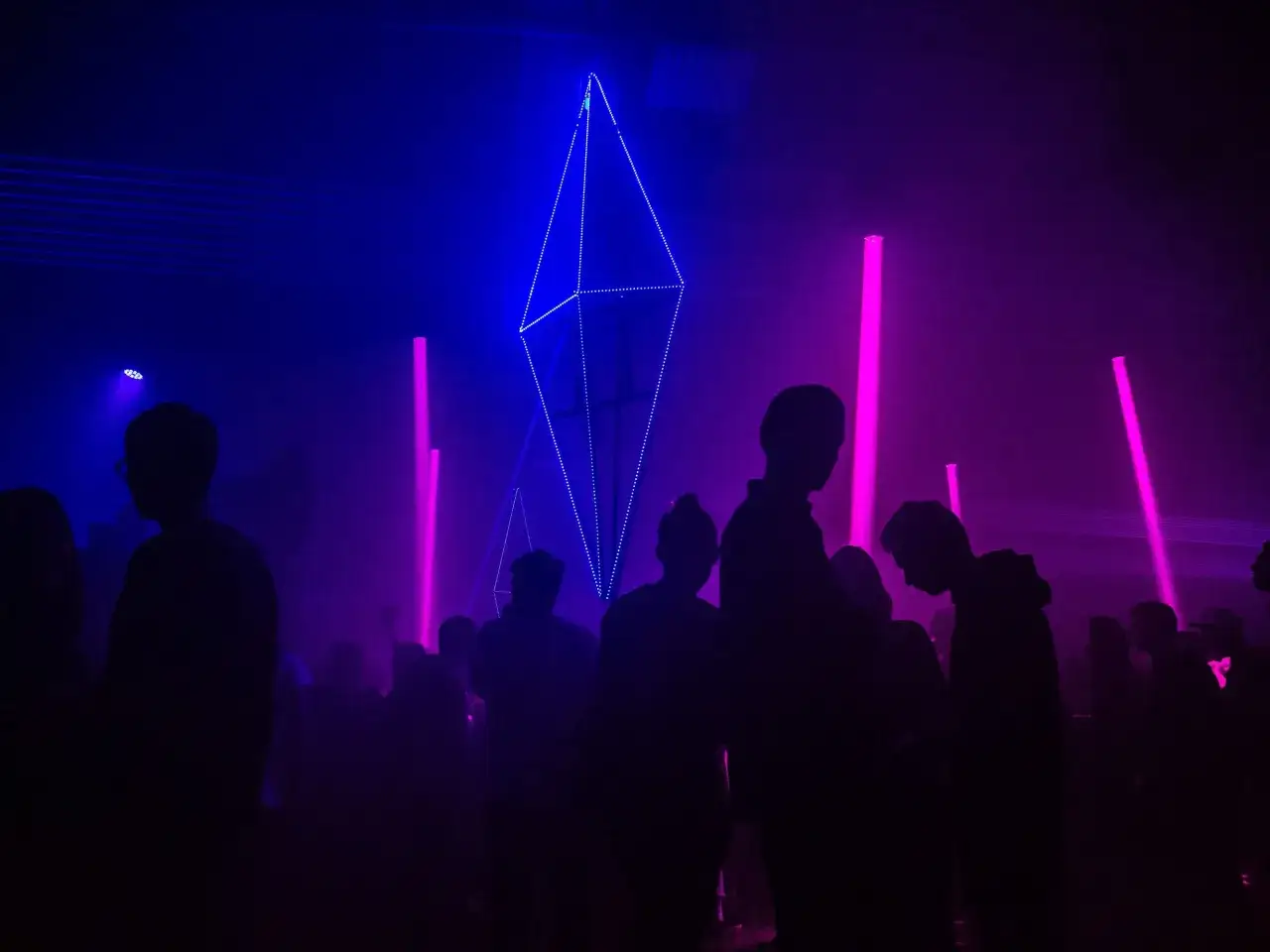 Silhouetten von Menschen tanzen im pulsierenden Licht einer Techno-Veranstaltung. Blaue und pinke Laserstrahlen durchschneiden den Nebel, w&auml;hrend eine geometrische Lichtinstallation im Hintergrund funkelt.