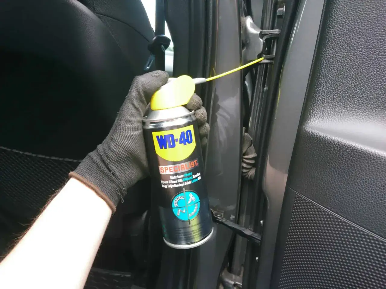 Ręka w rękawiczce aplikuje smar WD-40 na zawiasy drzwi, by wyeliminować skrzypiące drzwi.