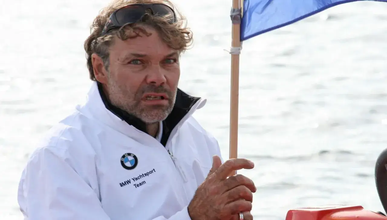 Markus Wieser, mit zerzaustem Haar und Sonnenbrille auf dem Kopf, tr&auml;gt eine wei&szlig;e BMW Yachtsport Team Jacke. Er h&auml;lt einen Holzstab mit einer blauen Flagge.