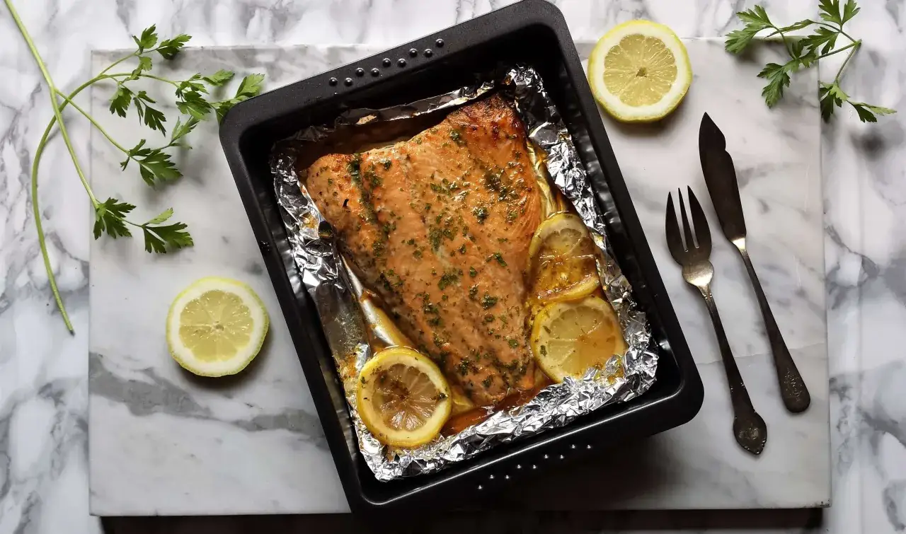 Un jugoso salm&oacute;n al horno, cubierto de hierbas y rodajas de lim&oacute;n, listo para ser servido con cubiertos de plata.