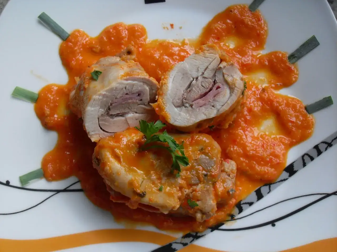 Contramuslos de pollo Thermomix enrollados con jamón, cubiertos de salsa de pimientos rojos y decorados con perejil y cebolleta.