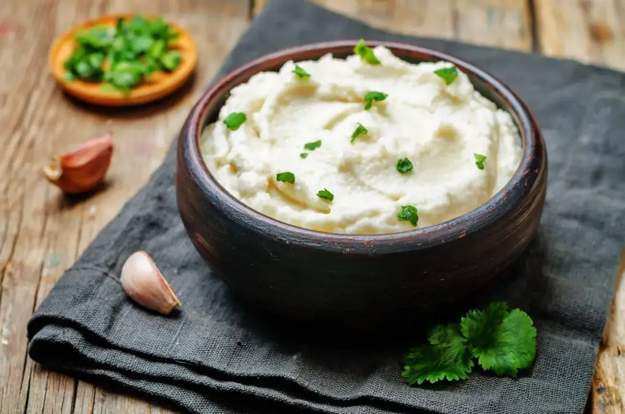 Aksamitne puree z kalafiora w glinianej misce, ozdobione natką pietruszki. Obok czosnek i mała miseczka z posiekanymi ziołami.