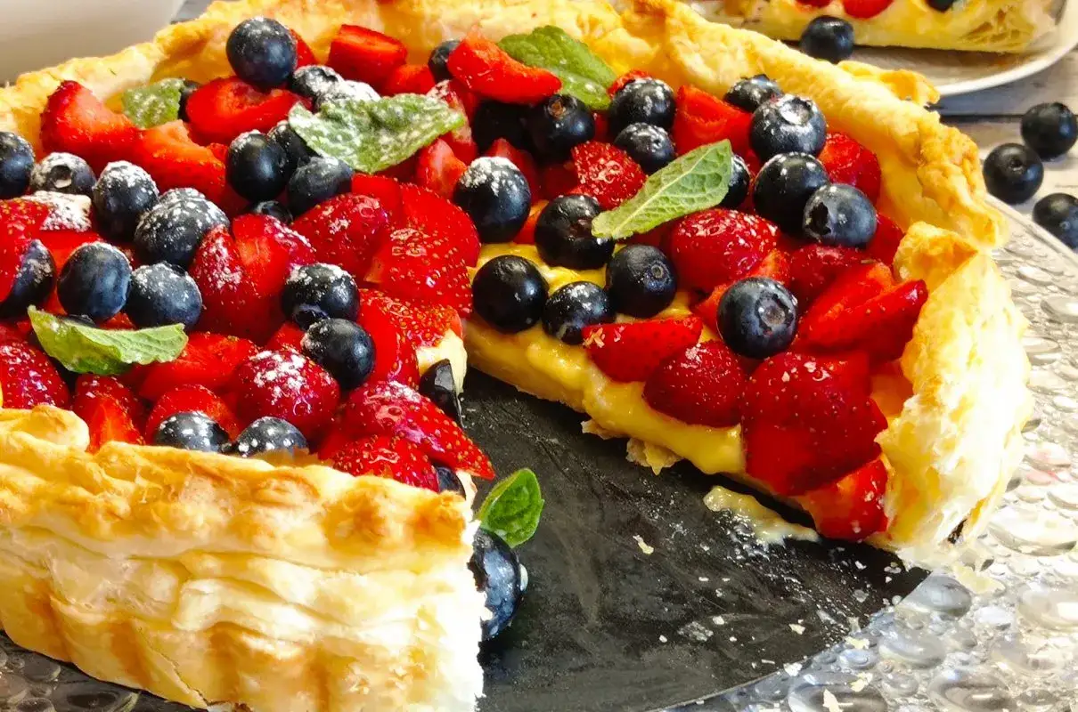 Pyszna tarta z ciasta francuskiego na słodko, obficie udekorowana truskawkami i borówkami, posypana cukrem pudrem.