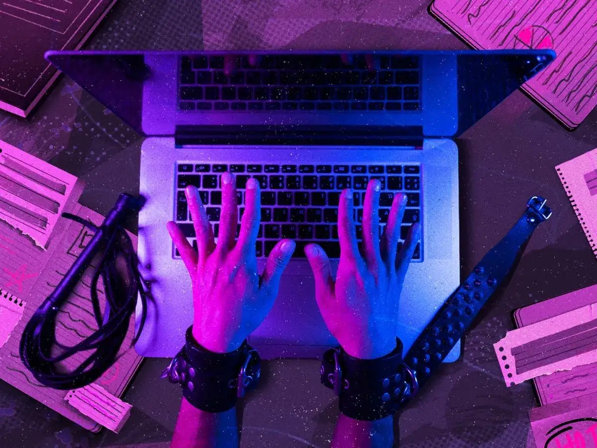 Ręce w kajdankach piszą na laptopie, obok bicz i notatki. To inspiracja do tworzenia treści na blog erotyczny.