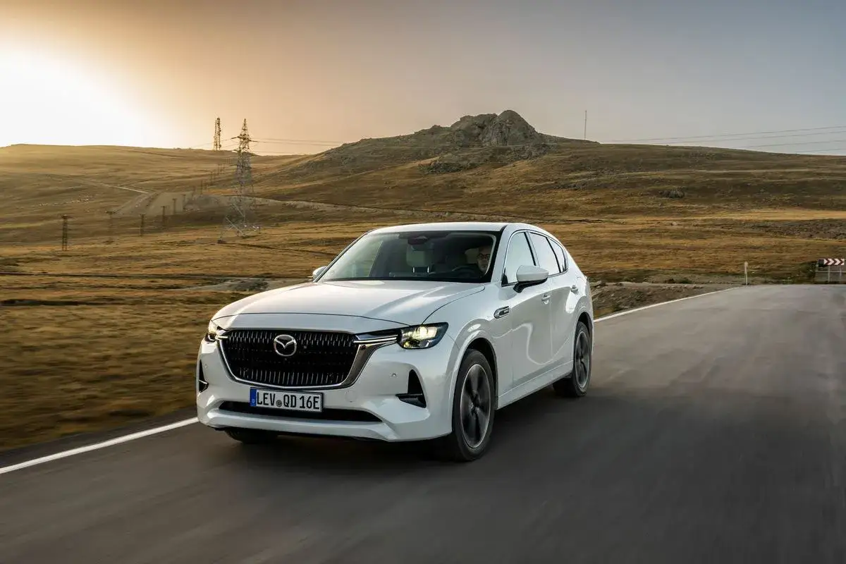 Vit Mazda CX-60 k&ouml;r p&aring; en v&auml;g i ett &ouml;kenlandskap. Kanske &auml;r det en **Mazda CX-60 problem** med att hitta en laddstation h&auml;r?