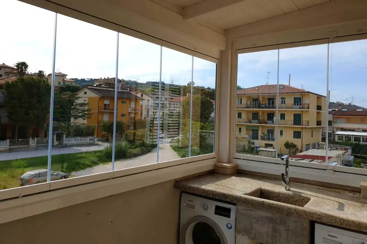 Un balcone trasformato con vetrate scorrevoli, un'idea per chiudere un balcone fai da te. Lavatrice e lavello in primo piano.
