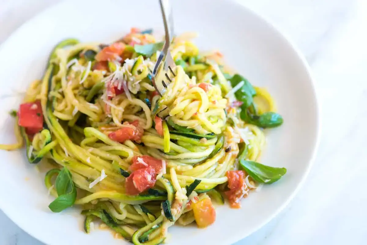 Eine Gabel hebt eine Portion leckerer Zucchini-Nudeln mit Tomaten und Basilikum von einem Teller.