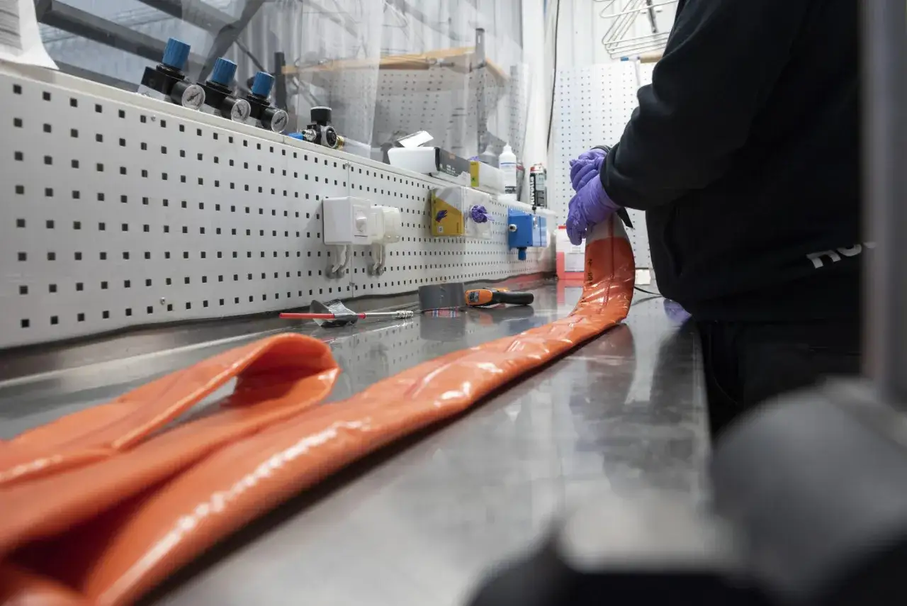 Person med lila handskar förbereder en orange slang för relining avlopp. Verktyg och material syns på arbetsbänken.