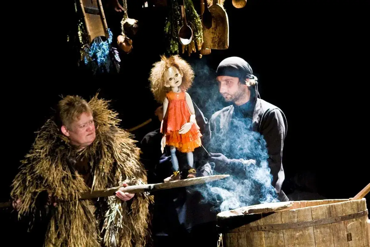 Baba Jaga patrzy, jak lalka w pomarańczowej sukience tańczy w dymie. Obok stoi mężczyzna w słomianym płaszczu.