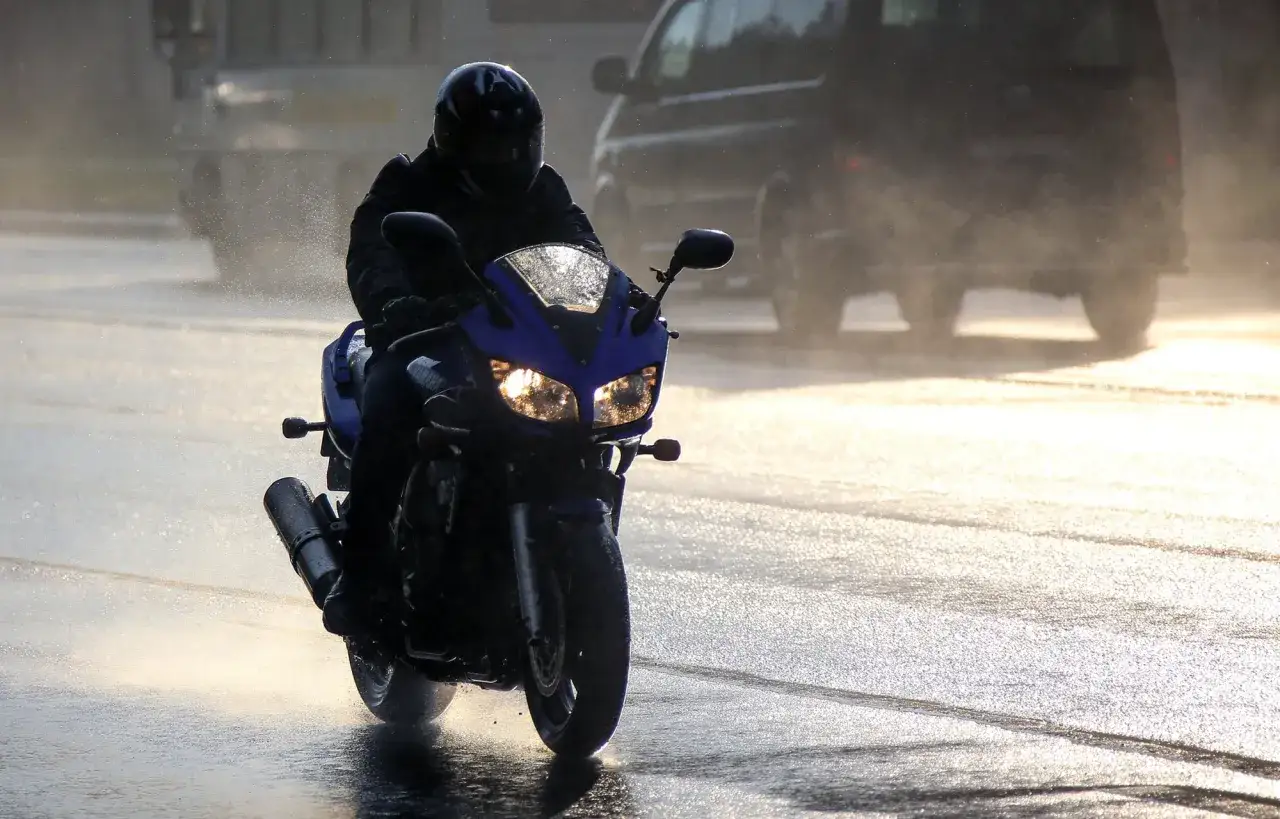 Motocyklista w kasku przemierza mokrą drogę. Jazda motocyklem w deszczu wymaga skupienia, a światła pojazdu odbijają się w kałużach.