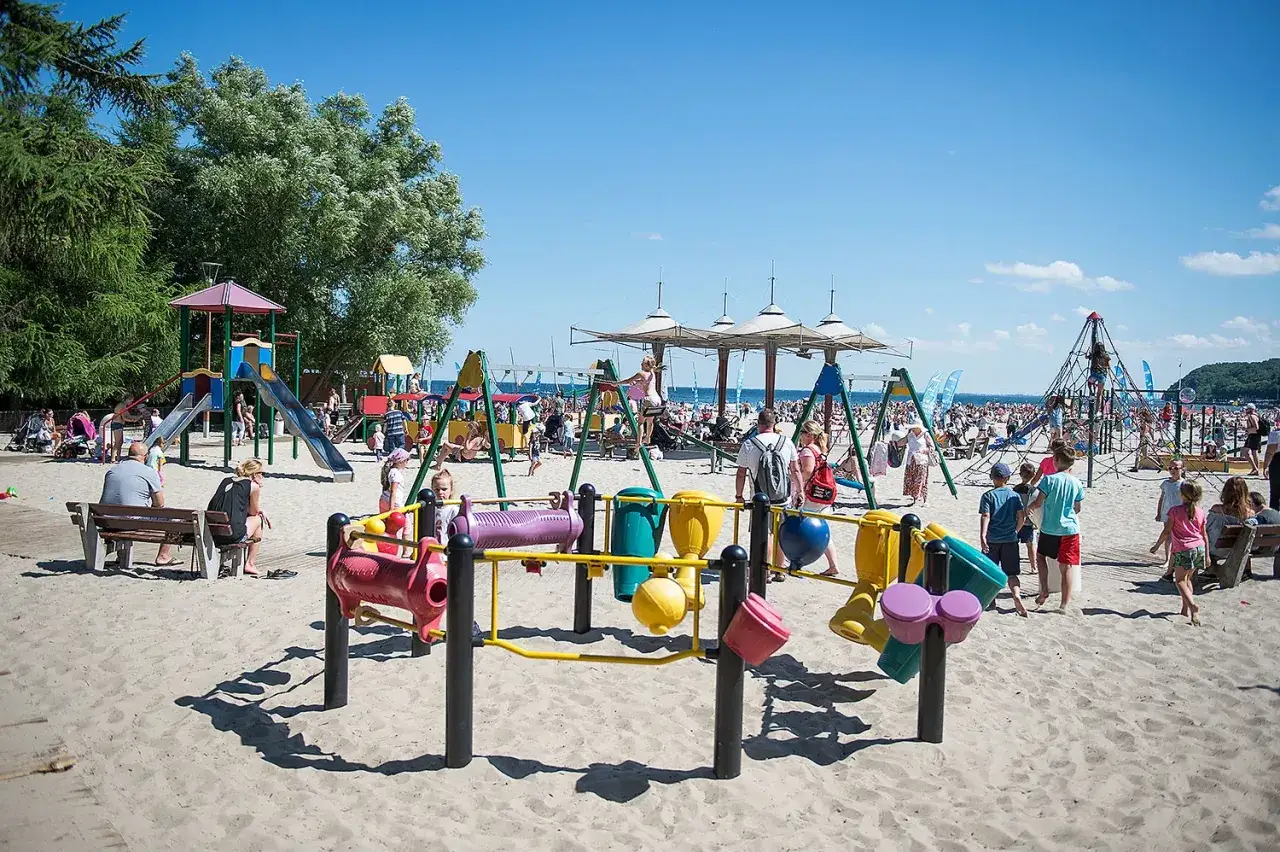 Kolorowy plac zabaw na plaży w Gdyni oferuje mn&oacute;stwo atrakcji dla dzieci. W tle widać tłumy plażowicz&oacute;w i morze.
