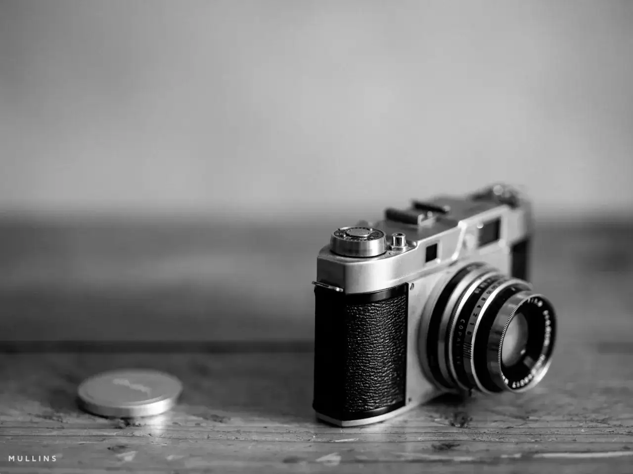 A vintage Voigtlander Bessa camera rests on a wooden surface, its silver body and textured grip catching the light. A lens cap sits nearby.
