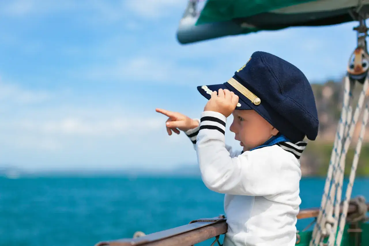 Bambino con cappello da capitano su una barca a vela, indica l'orizzonte con un dito.