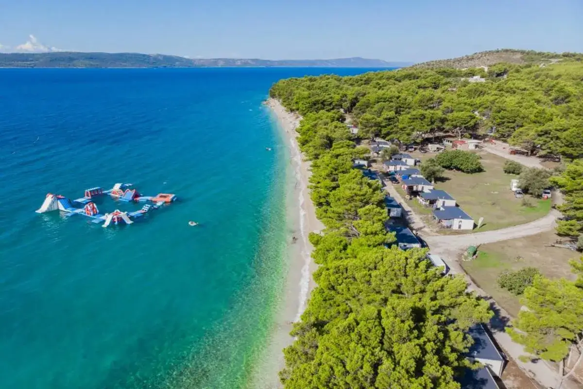 Wypoczynek w camp Dole Živogošće: wodny plac zabaw w turkusowym morzu, piaszczysta plaża i domki kempingowe wśród sosen.