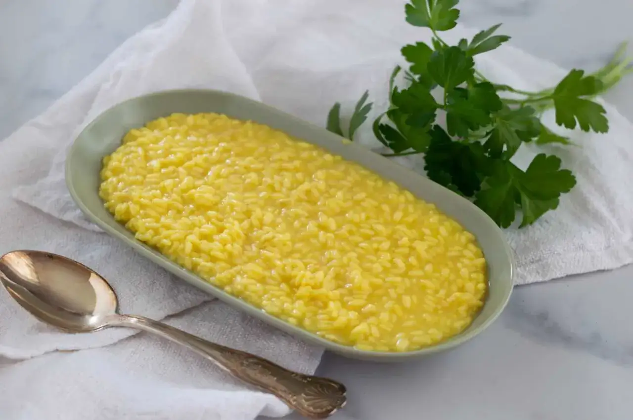 Un cremoso risotto alla milanese, giallo intenso, servito in un piatto ovale con un rametto di prezzemolo fresco e un cucchiaio d'argento.