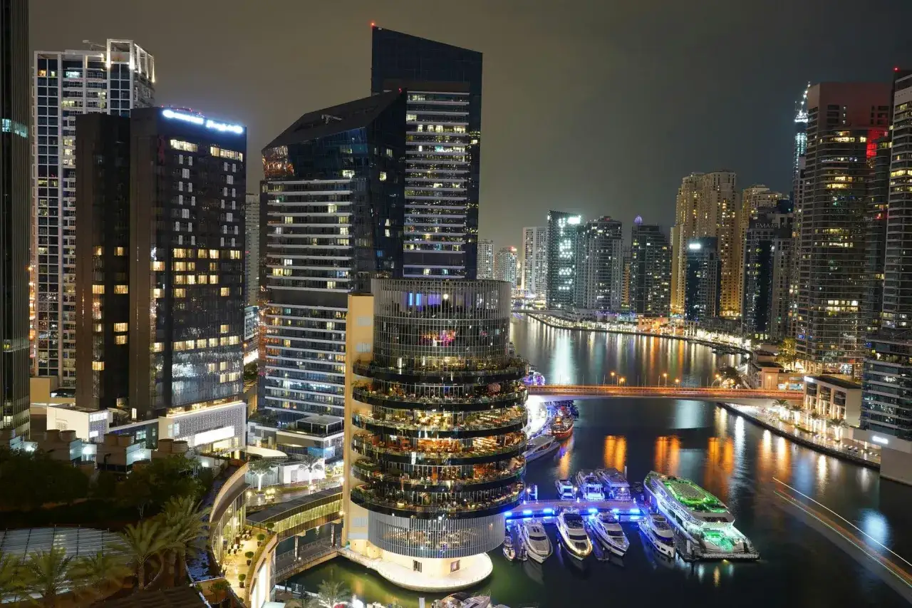 En glittrande stadssilhuett i Dubai i december. Höga byggnader lyser upp natten vid en kanal där lyxyachter ligger förtöjda.