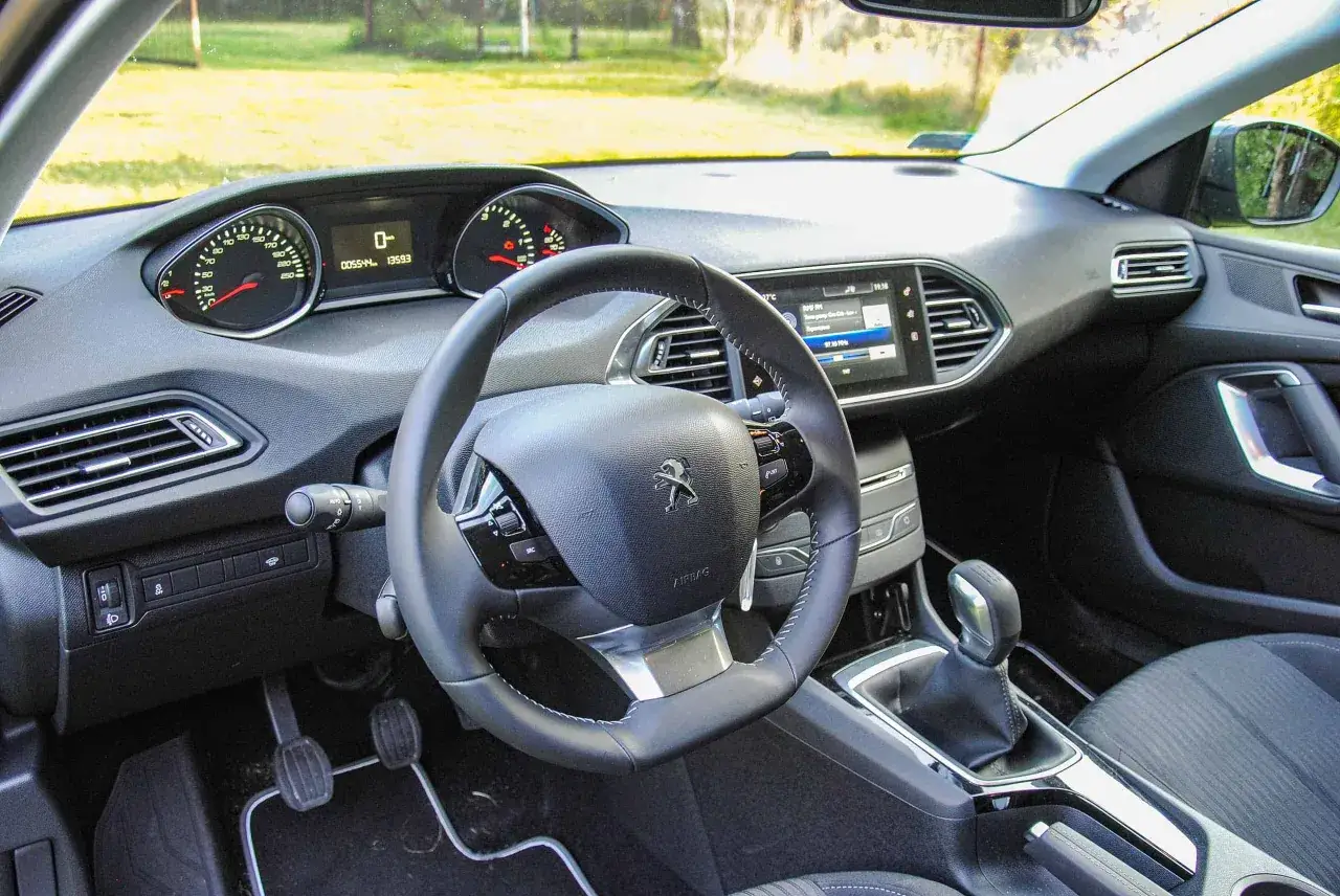Wnętrze Peugeot 308 T9: kierownica, cyfrowe zegary i ekran multimediów. Widoczne pedały i dźwignia zmiany biegów.