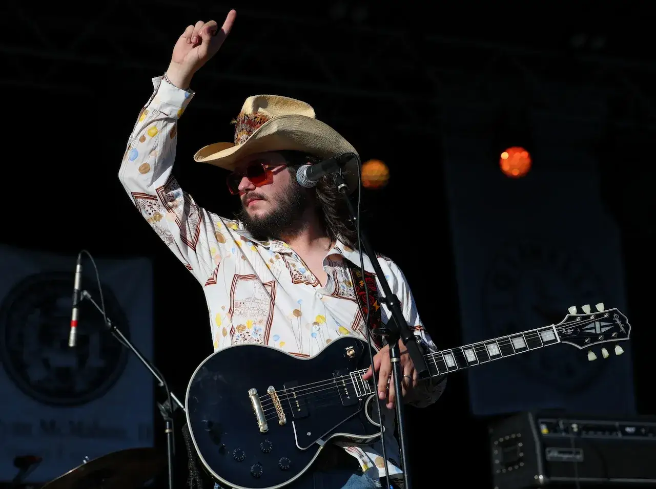 Un guitariste, chapeau de cowboy, chemise à motifs, lunettes de soleil, joue de la guitare électrique sur scène, l'air absorbé par le blues.
