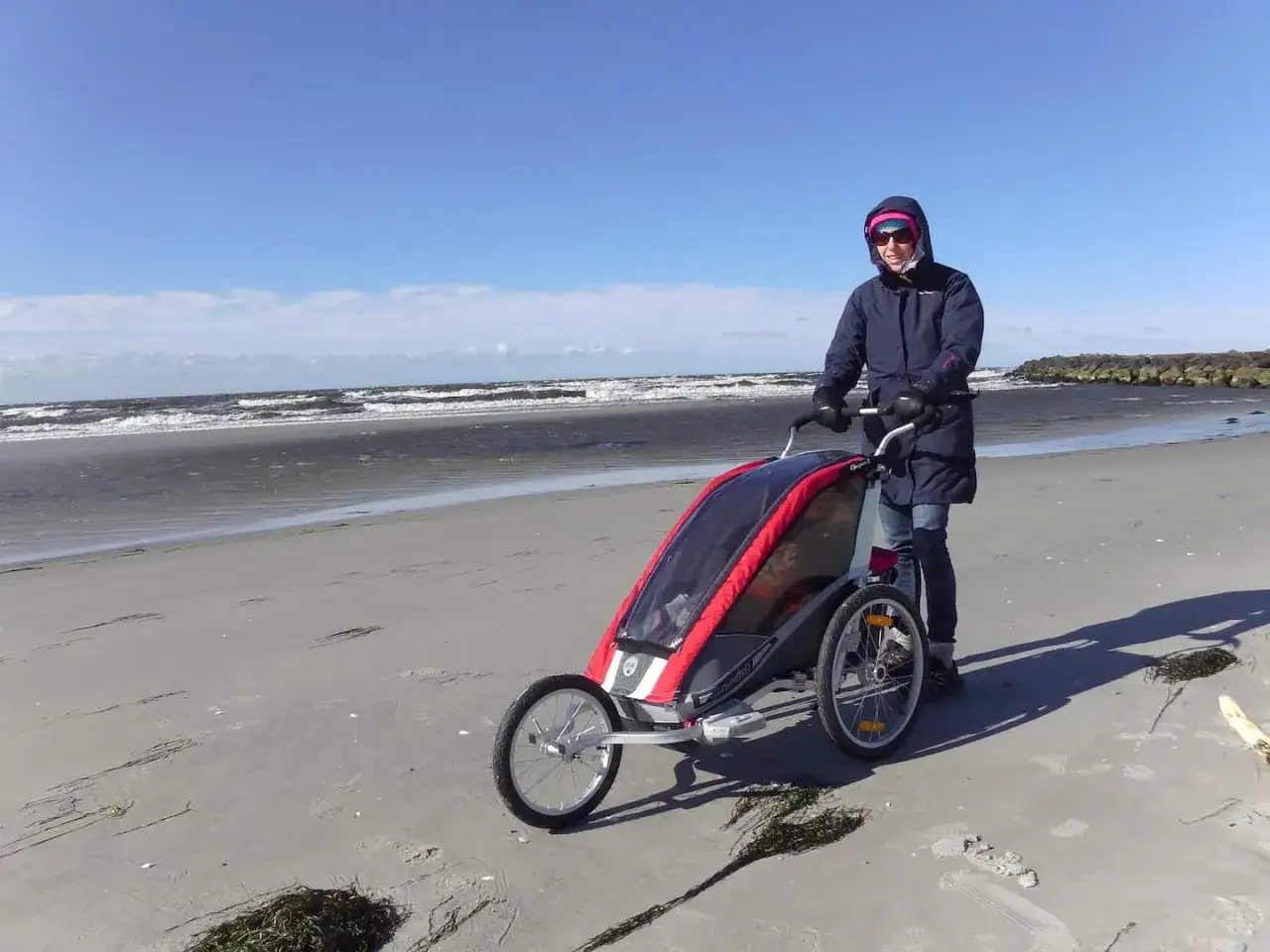 En person knuffar en multisportvagn på en sandstrand med havet i bakgrunden.