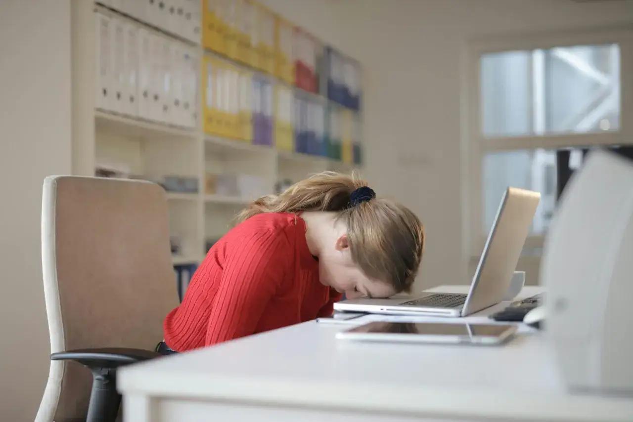 Zmęczona dziewczyna w czerwonym swetrze śpi na biurku, głowa oparta o laptopa. To obraz pracoholizmu.