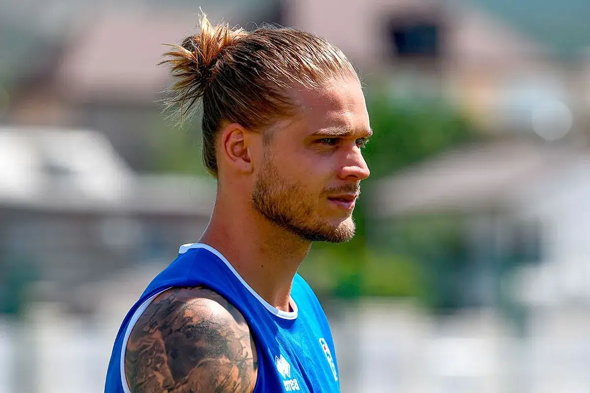 Mann mit man bun ohne bart, trägt blaues Trikot. Tätowierung am Arm.
