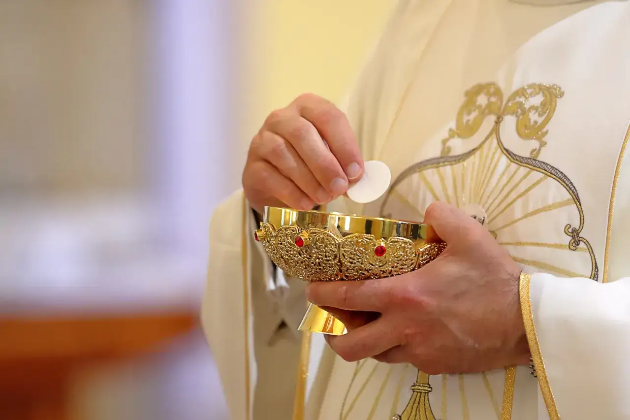Kapłan w białej szacie z haftem, trzymający złoty kielich i hostię. Symbolika sakramentu, nie podatek na kościół.