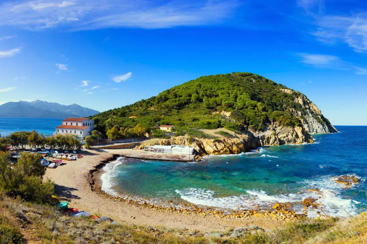 Baia incantevole all'Isola d'Elba, Toscana. Un promontorio verdeggiante si tuffa nel mare cristallino, con una spiaggia di ciottoli e barche colorate.