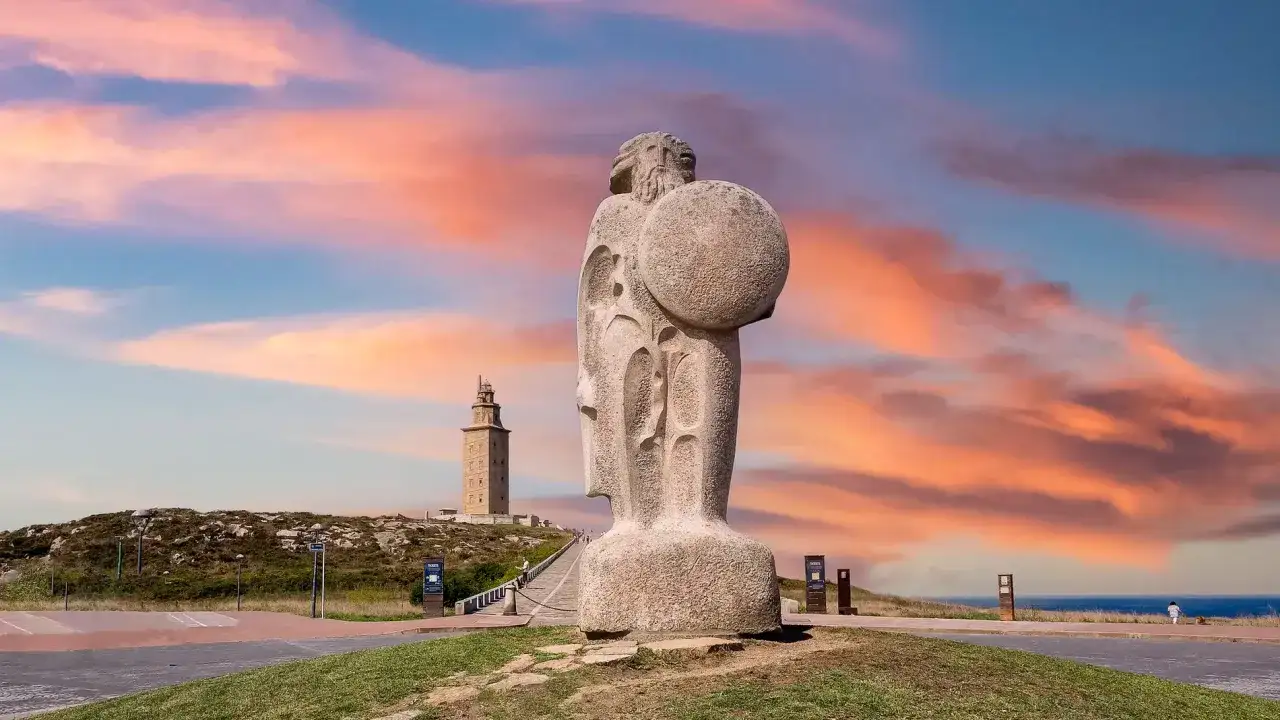 Gigantesca estatua de Hércules con escudo, uno de los monumentos de Galicia, ante la Torre de Hércules al atardecer.