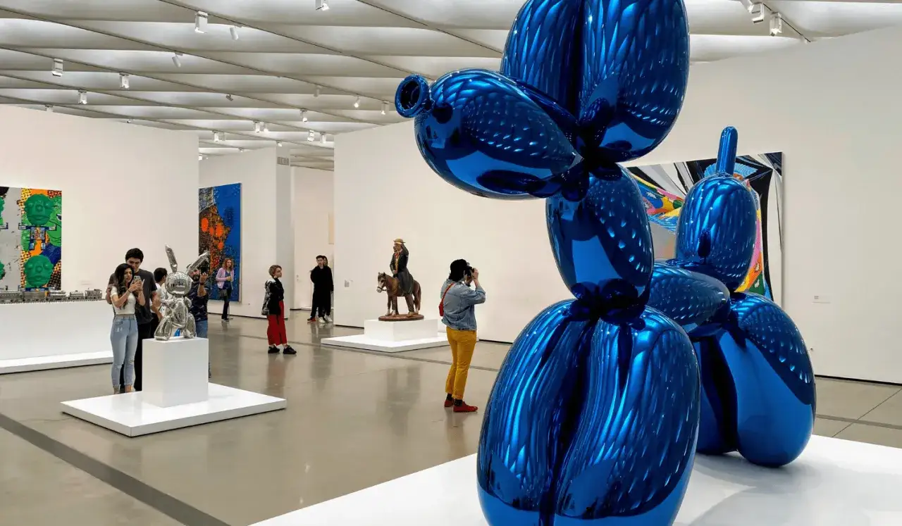 A blue balloon dog sculpture dominates the foreground, with visitors admiring art, perhaps aspiring billionaire art collectors, in a modern gallery.