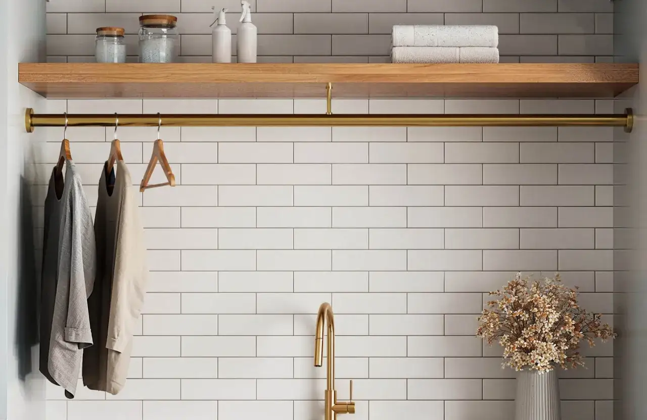 A laundry room with white tile and beige grout, featuring a wooden shelf with jars and towels, a brass rod with hanging clothes, and a brass faucet.