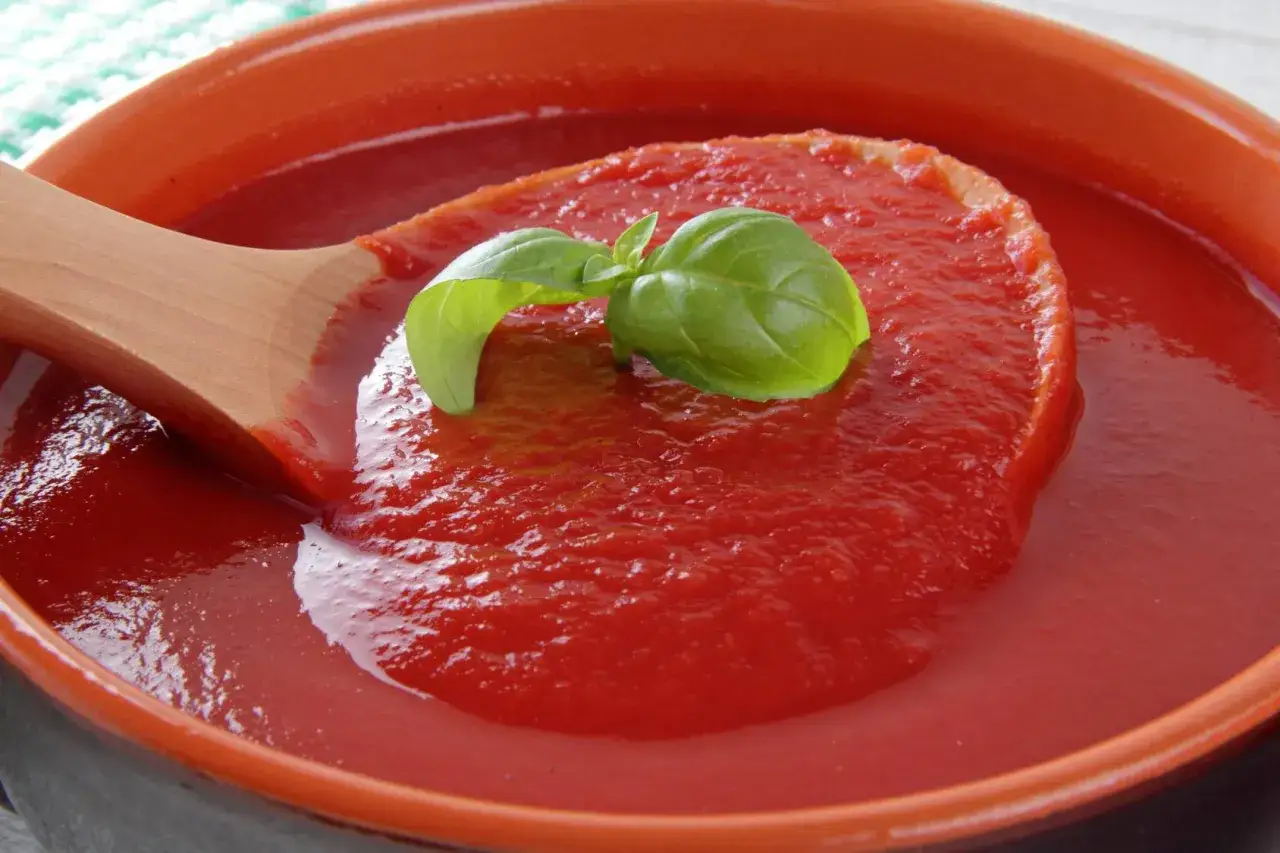 Gęsty, domowy ketchup w misce, nabierany drewnianą łyżką, ozdobiony listkiem bazylii. Idealny przepis na ketchup!
