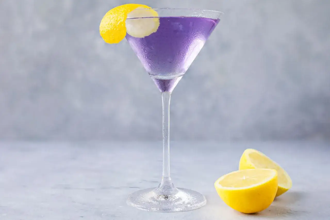 En lila aviation drink i ett martiniglas, garnerad med citronskal och en citronskiva. Två halvor av en citron ligger bredvid.