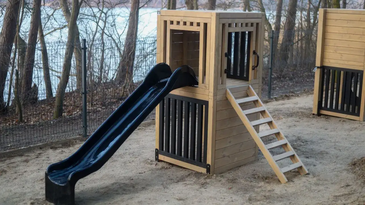 Drewniany plac zabaw ze zjeżdżalnią i drabinką, idealny na **camping dla dzieci**. Otoczony siatką, z widokiem na las i jezioro.