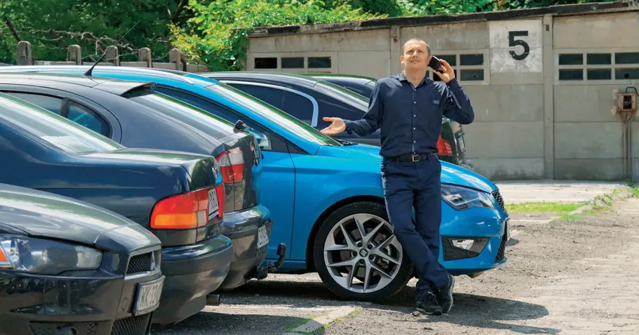 Mężczyzna rozmawia przez telefon, stojąc obok niebieskiego auta. Podpowiada, na co zwracać uwagę przy zakupie auta, analizując dostępne modele.