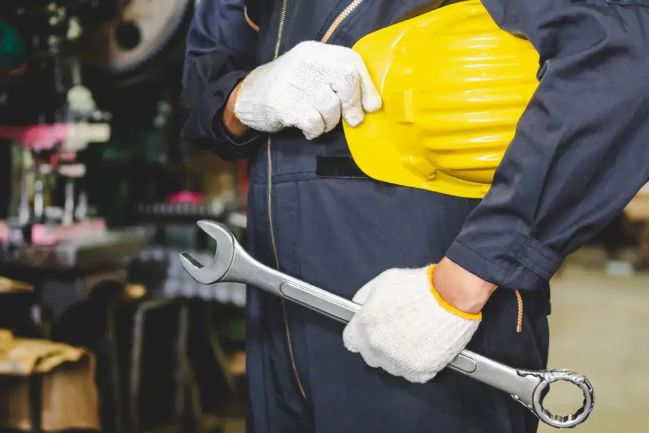 Un trabajador con guantes blancos sostiene una llave inglesa y un casco amarillo. Los epis son una medida de protección esencial para su seguridad en el taller.