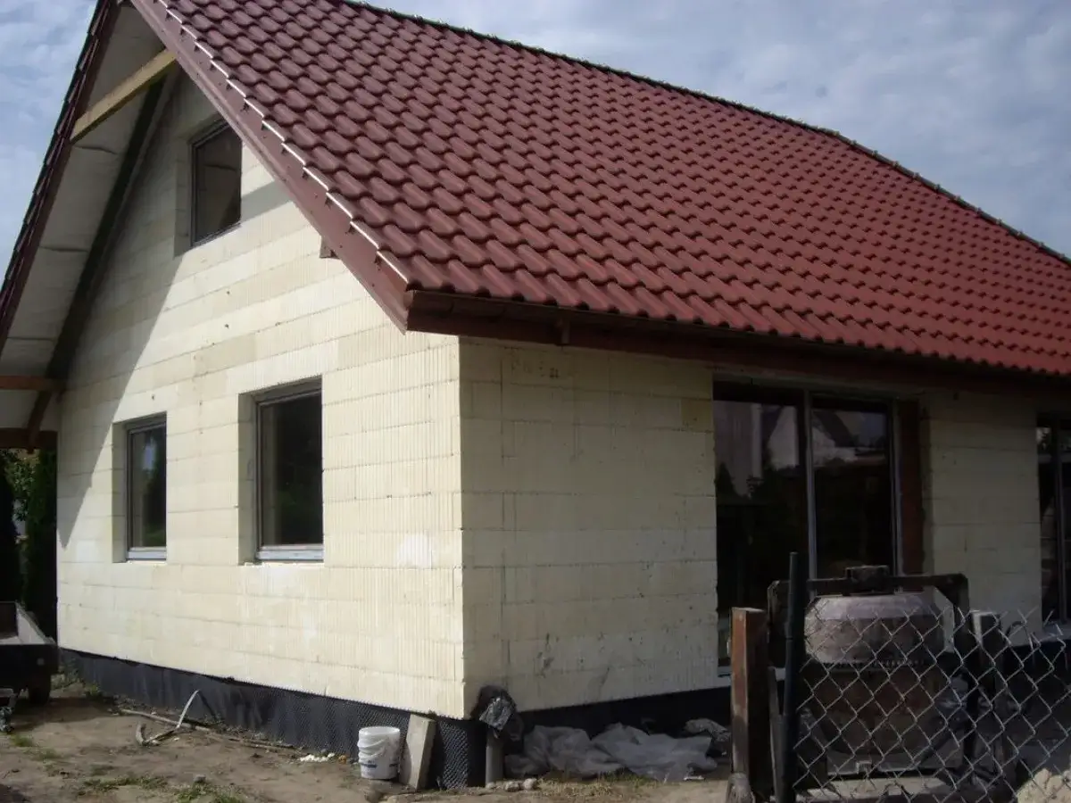 Budowa domu ze styropianu w toku. Jasne ściany, czerwony dach, okna i betoniera na placu budowy.