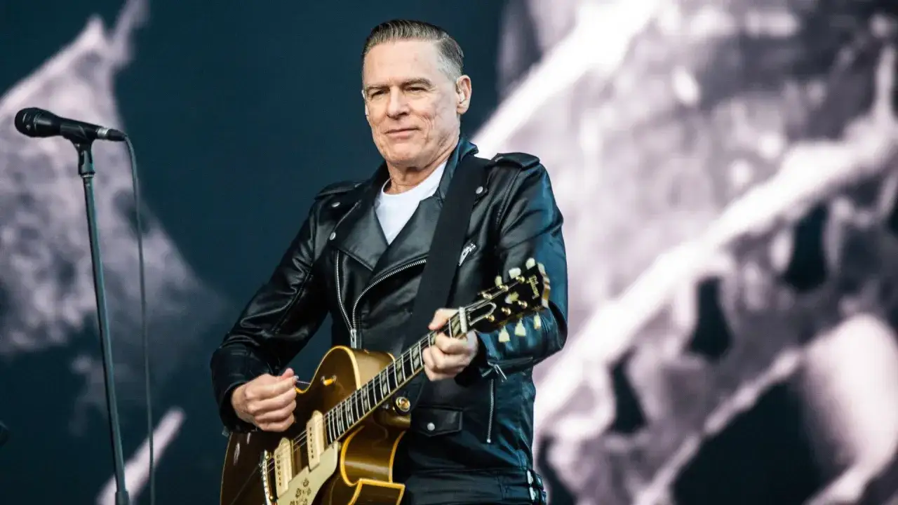 Bryan Adams w sk&oacute;rzanej kurtce gra na gitarze, przypominając swoje kultowe utwory.