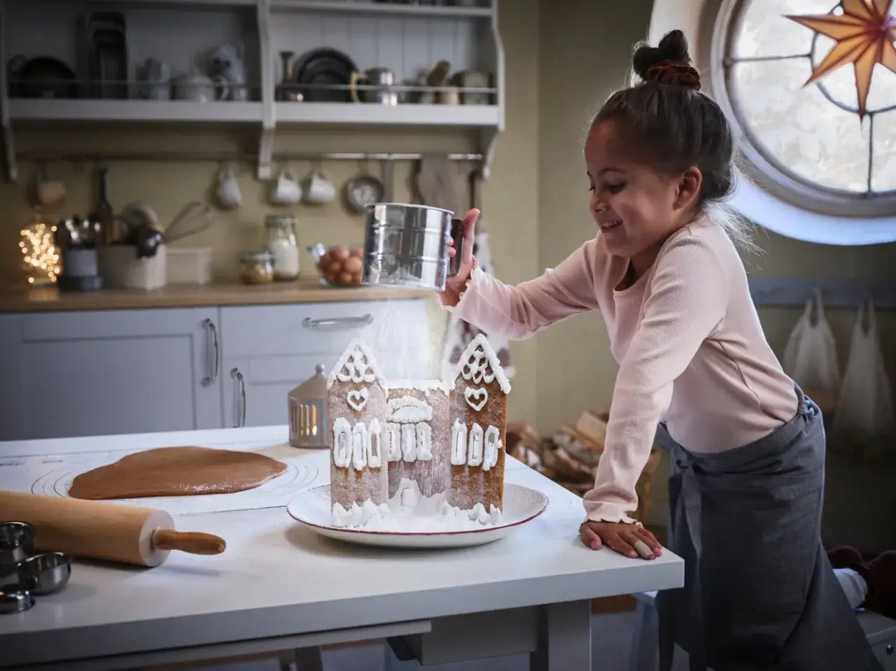 Glad flicka pudrar florsocker över pepparkakshus. Ett mysigt julbaka med barn i köket.