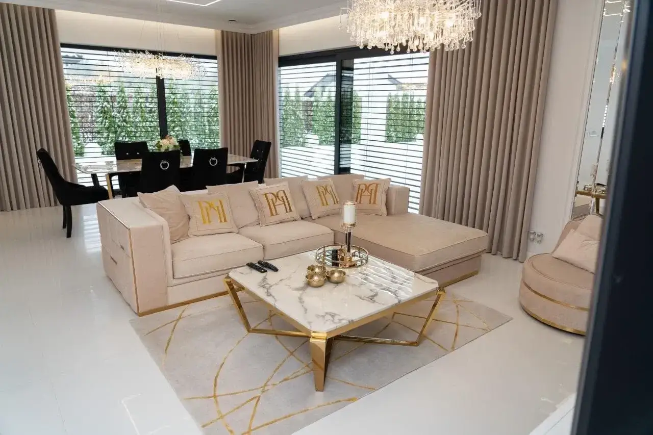 Elegancki salon glamour z beżową sofą, marmurowym stolikiem i złotymi akcentami. W tle jadalnia z czarnymi krzesłami.
