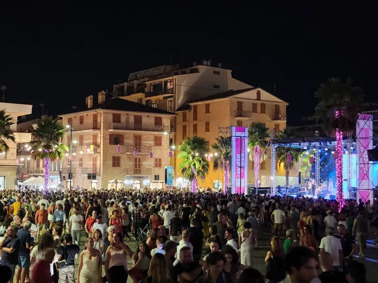Folla festante a Porto Sant'Elpidio per la Notte Rosa, con musica dal vivo e palme illuminate.