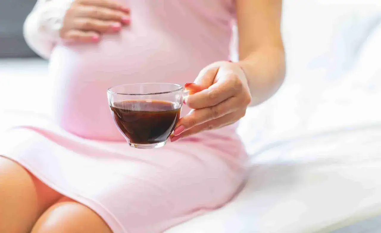 Mujer embarazada en vestido rosa sostiene una taza de té oscuro.