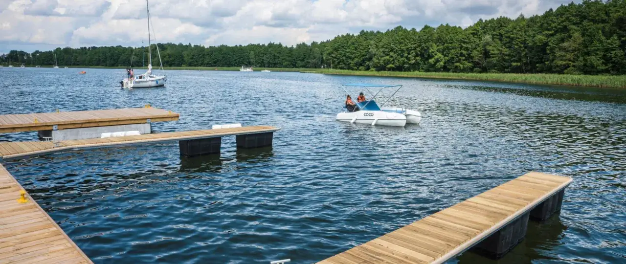 Drewniane pomosty na jeziorze, idealne miejsce, by zapytać: gdzie na Mazury? W oddali żaglówka i rower wodny 