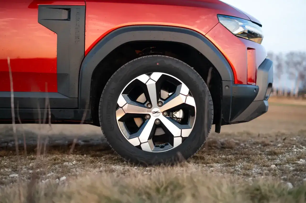 Czerwona Dacia Duster hybryda na tle suchej trawy. Widoczna felga z charakterystycznym wzorem i logo marki.