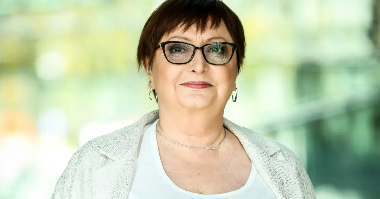 Krystyna Tkacz w okularach, w białej bluzce i marynarce. W tle rozmyte zielenie, jakby wspomnienie dzieciństwa.