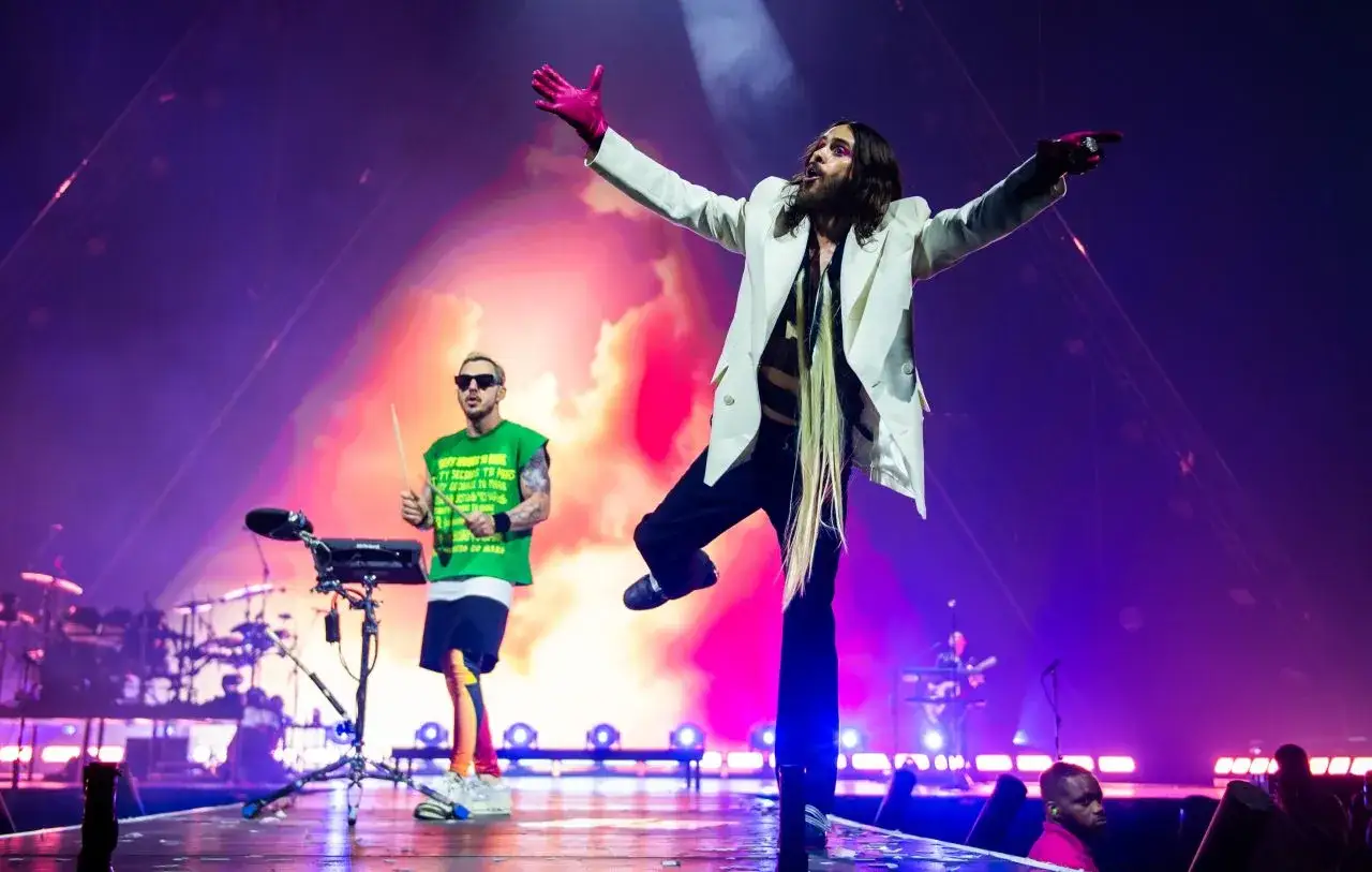Jared Leto, membre de Thirty Seconds to Mars, en veste blanche et gants roses, danse sur scène. À gauche, un autre membre joue de la batterie.