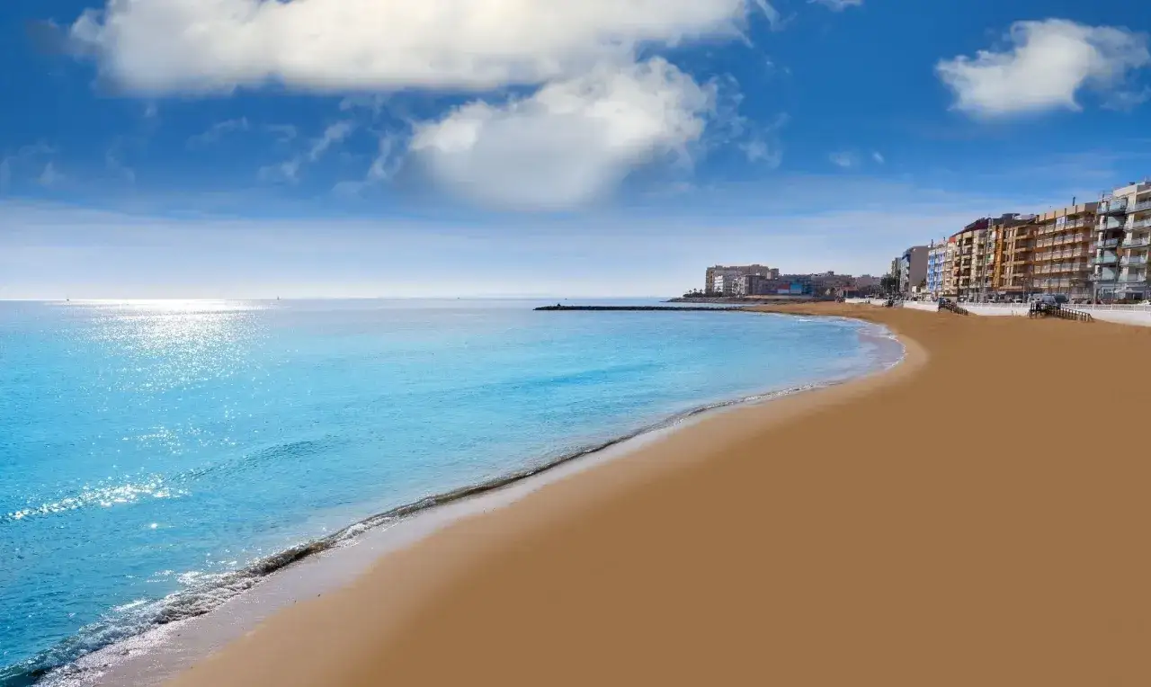 La **playa de los locos** se extiende dorada bajo un cielo azul con nubes algodonosas. El mar turquesa acaricia la arena, mientras edificios modernos se alinean en el horizonte.