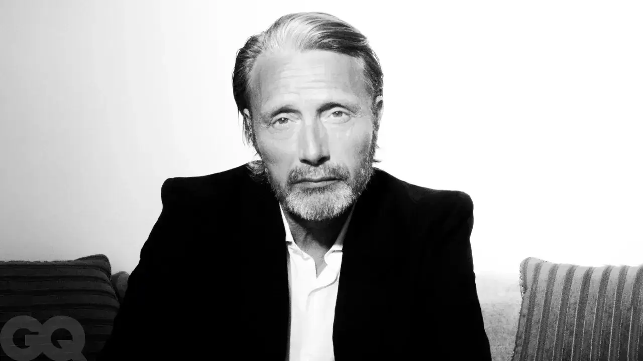 Mads Mikkelsen, med gråsprängt hår och skägg, bär en svart kavaj och vit skjorta. En GQ-logotyp syns i nedre vänstra hörnet.